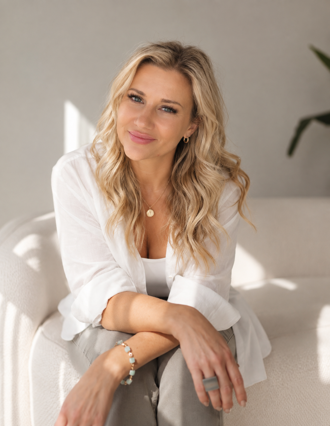 Krysten sitting on a cream sofa in a white blouse, smiling